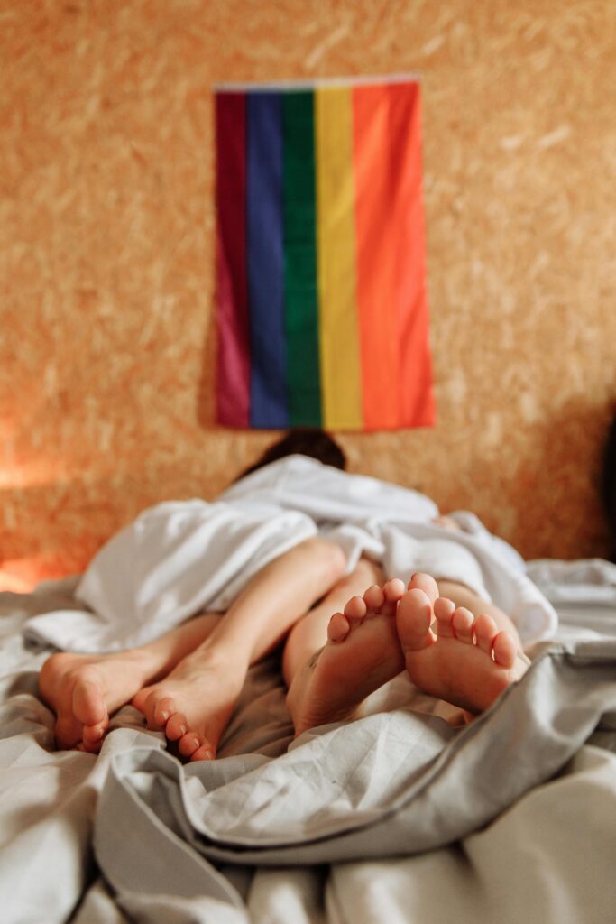 Lesbian couple lying together under a blanket with a rainbow flag on the wall, symbolizing LGBTQ+ pride.