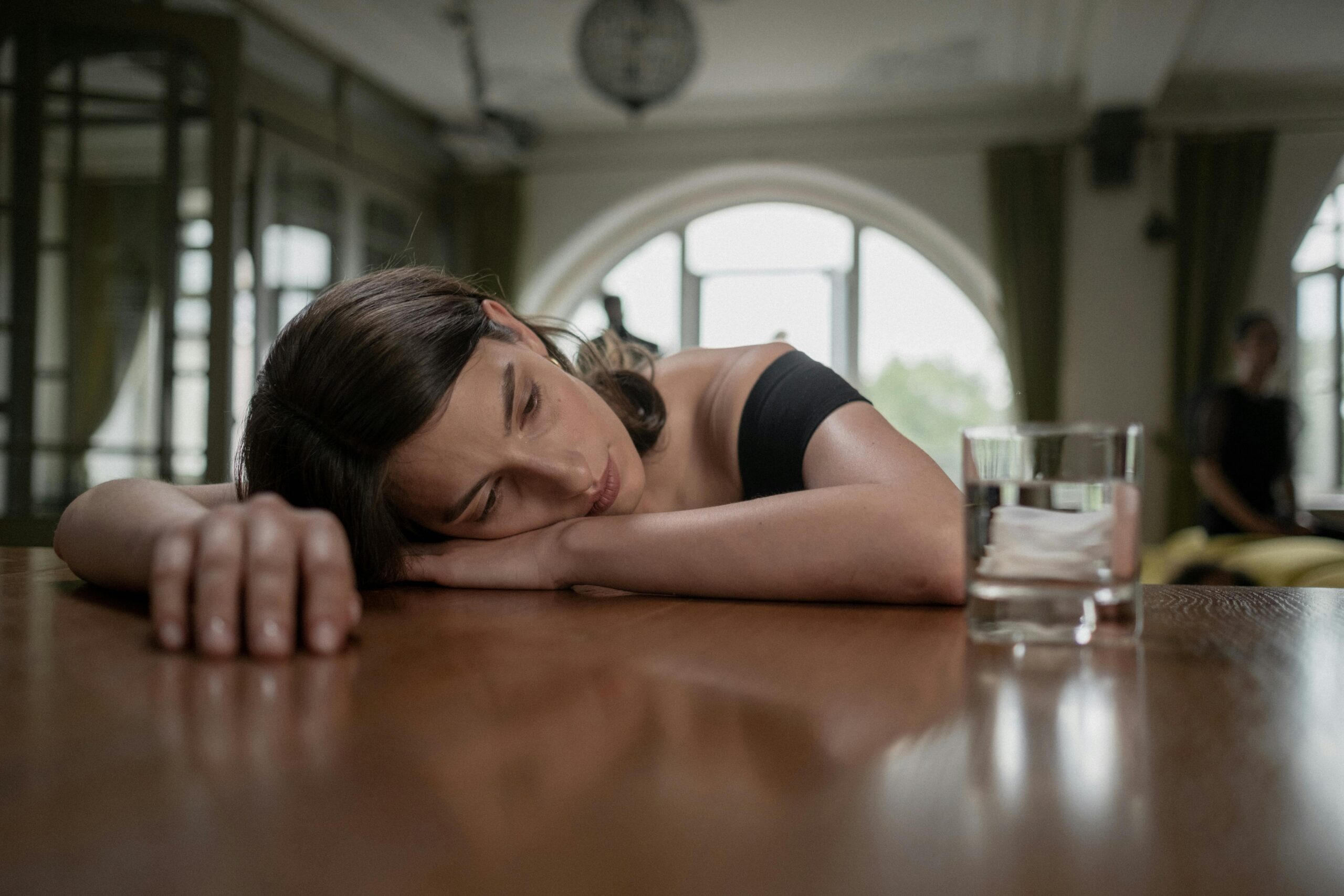 A woman in a black dress rests her head on a table beside a glass of water, conveying sadness.