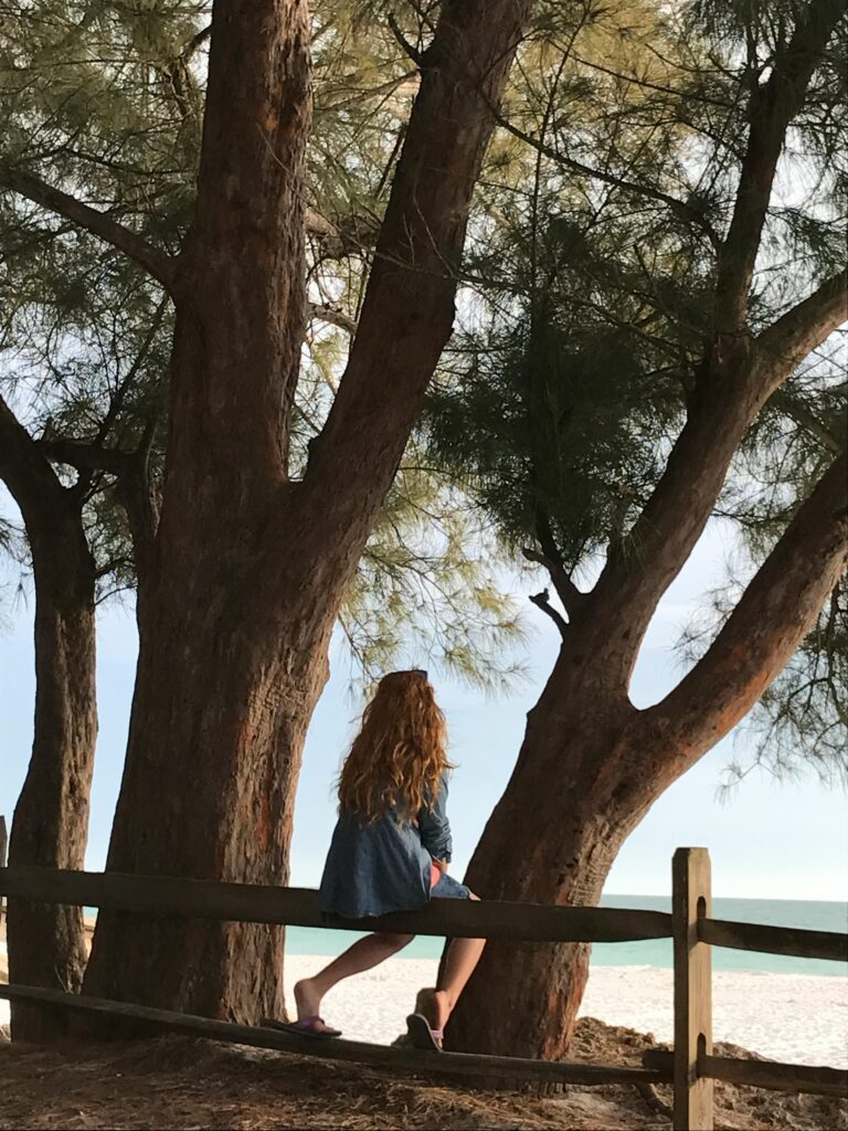 Woman with long blonde hair sitting by trees on a sandy beach, enjoying solitude.