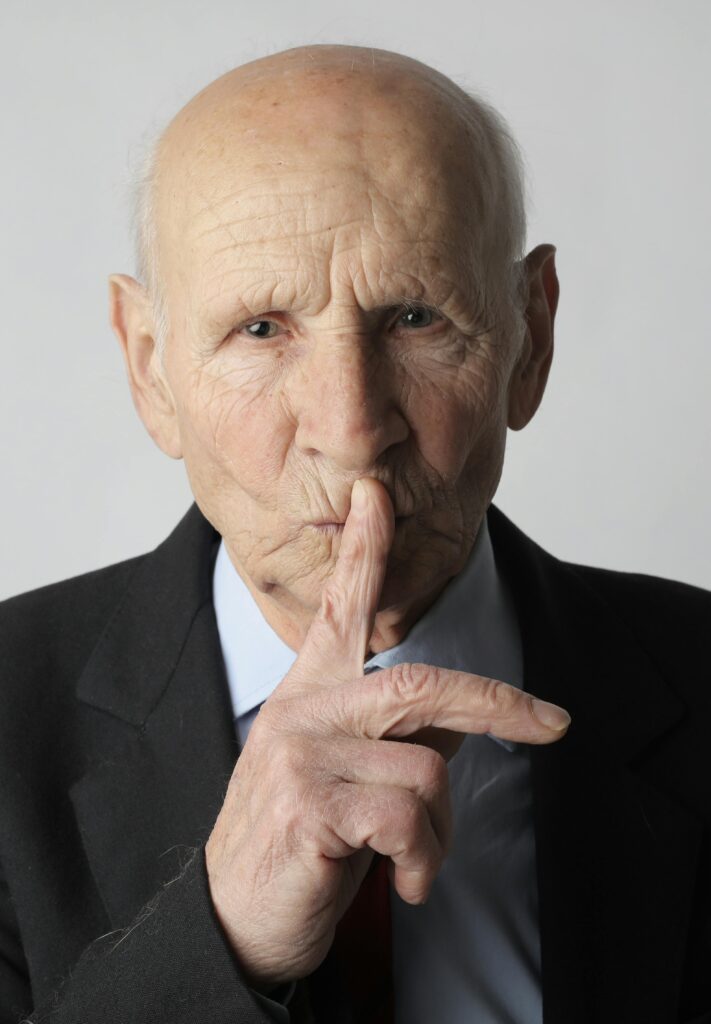 Portrait of a senior man wearing a suit, gesturing for silence in a studio setting.