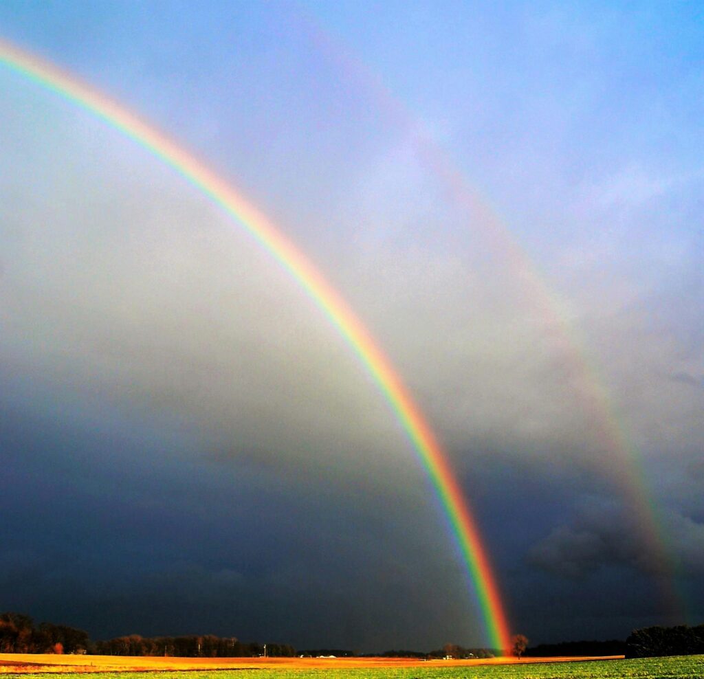 Double rainbow arching over a serene rural field against a dramatic sky.