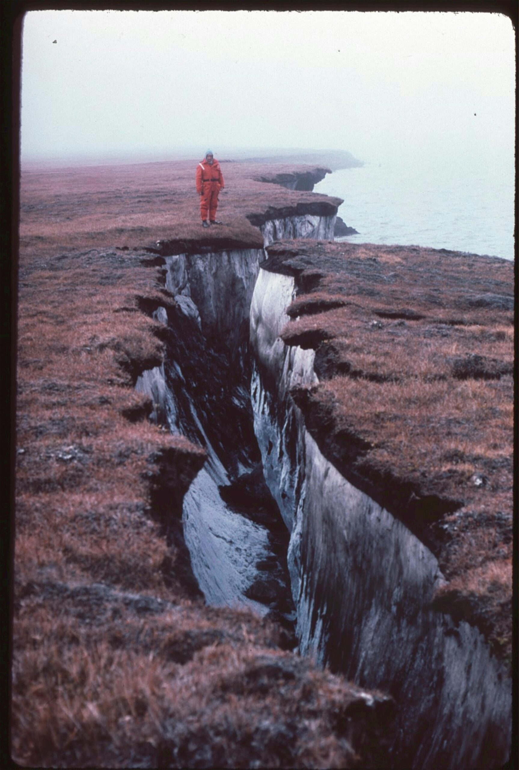 A lone person stands near a deep cliff crevice by the eroded seacoast.