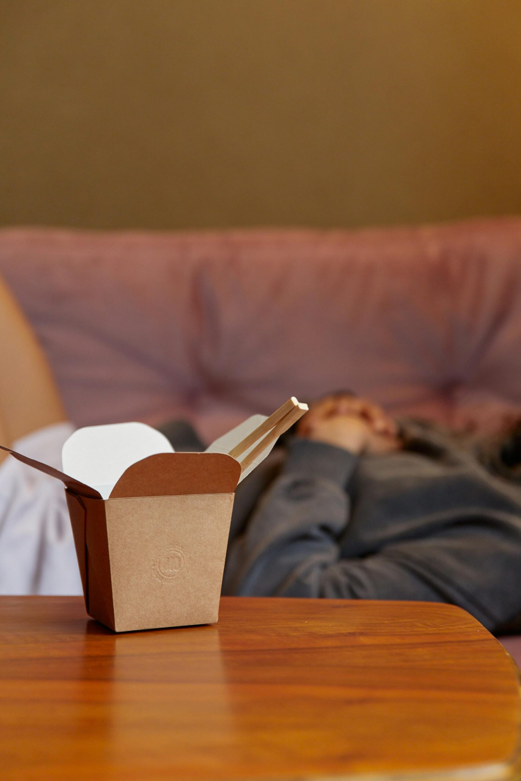 Brown takeout box on wooden table with blurred background of a person resting.