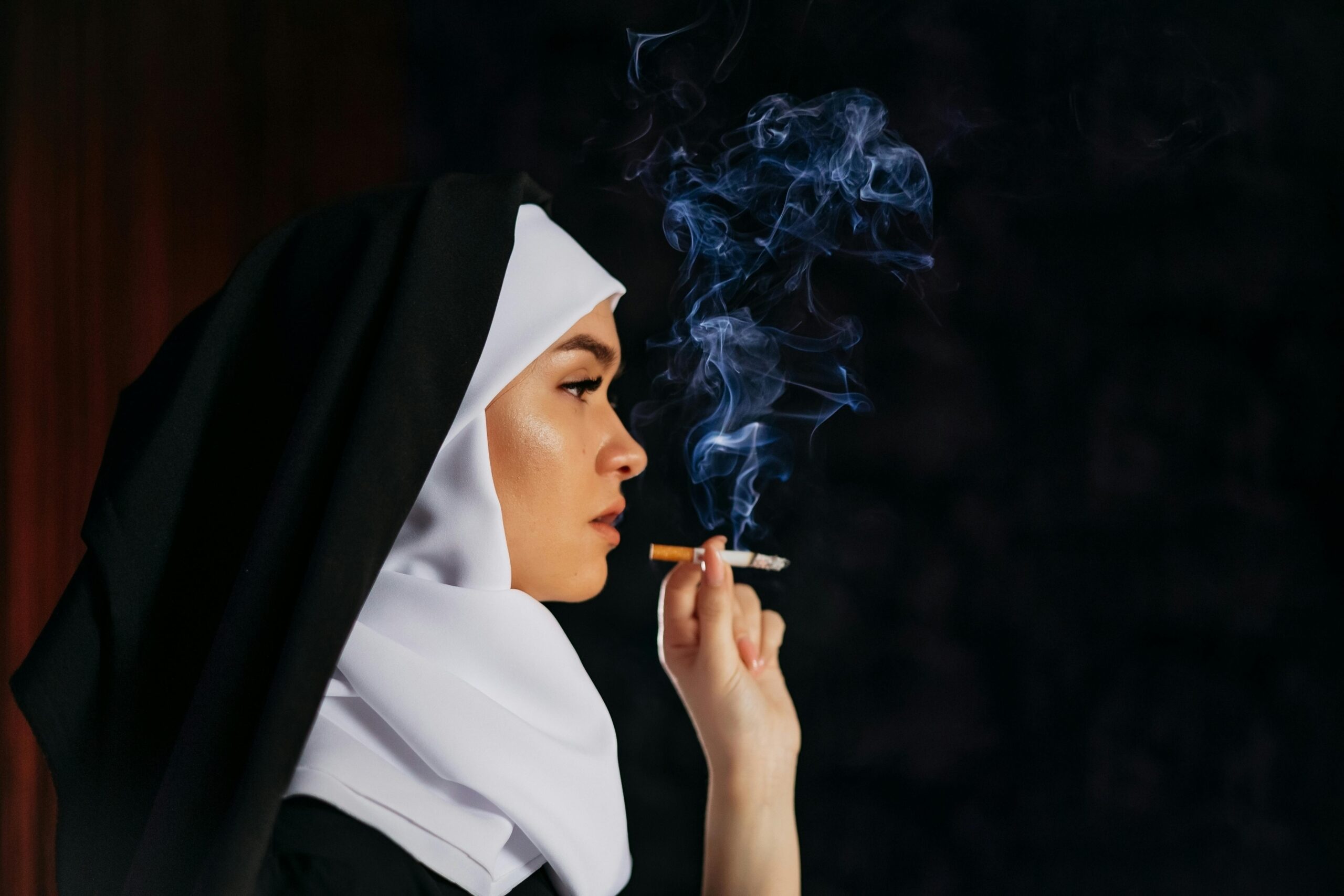 Profile of a nun in traditional habit smoking a cigarette, with smoke swirling against a dark backdrop.
