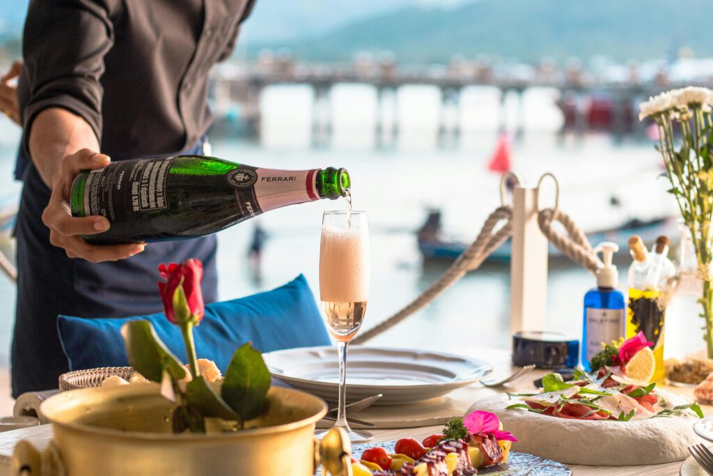 A waiter pours champagne at an elegant outdoor dining setup by the sea, creating a luxurious atmosphere.