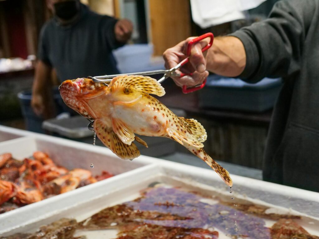 A vendor holds a fresh fish with tongs at a bustling seafood market.